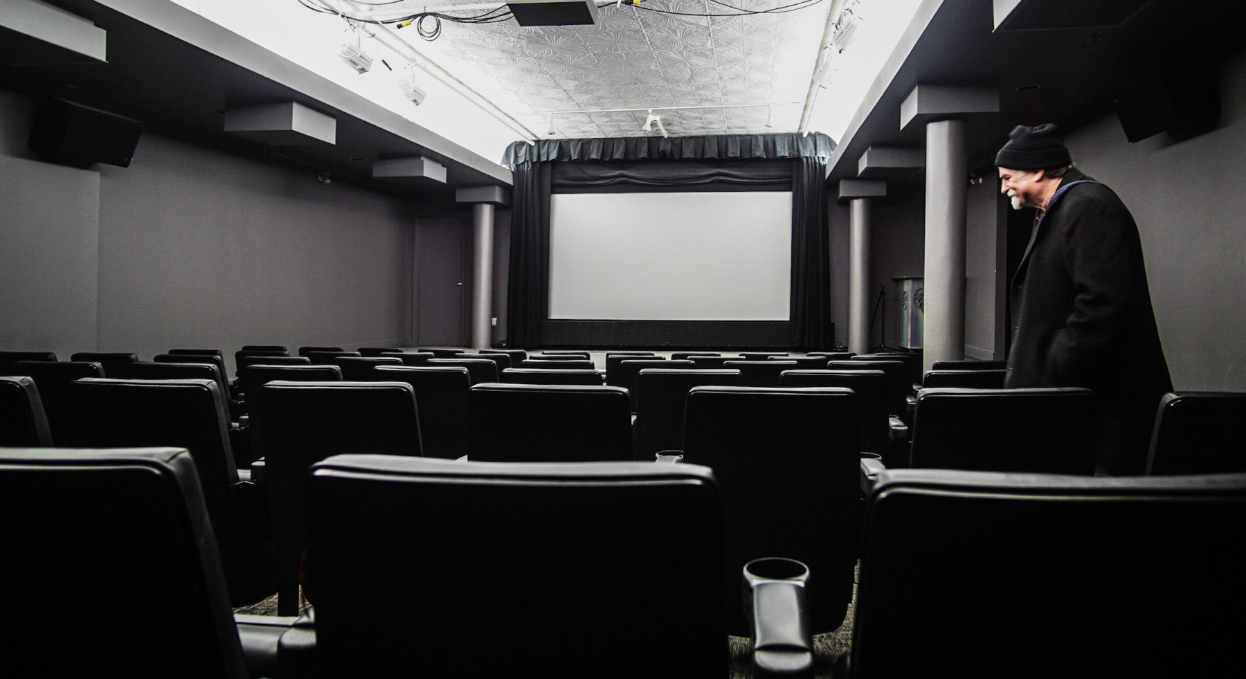 Dave Barber, a white man in his 60s in a beanie and winter coat walking past rows of empty seats in a small cinema, the Dave Barber Cinematheque with a blank screen.